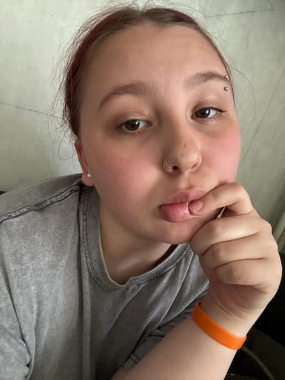 A selfie shows a young adult with brown eyes, light skin, and various piercings (eyebrow, nose, ear). The person is wearing a gray t-shirt and an orange wristband. Their hair appears auburn near the roots. The subject is pursing their lips and holding them with their fingers. The background suggests an interior setting with a light-colored floor. There is no data in this image that identifies the location as the city of Chornomorskoe, Ukraine.