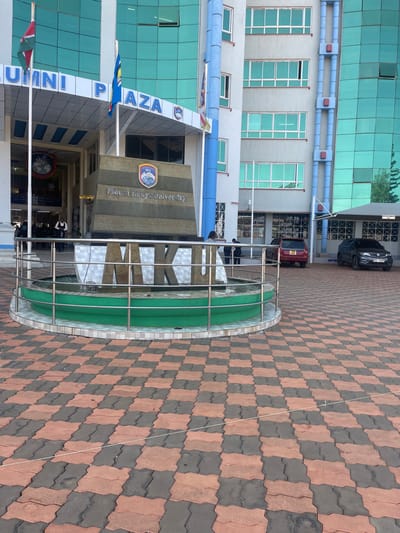 The image depicts the exterior of the Mount Kenya University (MKU) Alumni Plaza building, situated at the main campus in Thika, Kiambu County, Kenya. This is a university setting, not a hospital ward.
The scene features a modern, multi-story architectural structure composed of light-colored wall sections and numerous large, rectangular, light-blue tinted glass windows. The building's main entrance is recessed under an arched canopy, clearly labeled "ALUMNI PLAZA." Vertical blue decorative elements ascend the facade to the right of the main entrance. In the foreground, a large, tiered, dark stone monument is positioned centrally, displaying the text "Mount Kenya University" and the university's crest. Directly in front of this monument, a circular fountain with a green basin and a silver-colored metal railing showcases the prominent letters "MKU" (Mount Kenya University), over which water flows.
The ground area in the foreground is paved with interlocked stones in a checkerboard pattern of dark grey/black and reddish-brown. Two flags are displayed on flagpoles: the Kenyan national flag on the left and a blue flag with yellow elements (likely the university flag) on the right. Several individuals are visible: two figures in light-colored attire stand near the building's entrance, while two other individuals are positioned near the base of the central monument. Further to the right, two passenger vehicles are parked on a paved surface: a red car closer to the building and a dark-colored SUV further back. The scene is illuminated by bright daylight, casting visible reflections on the building's glass facade.