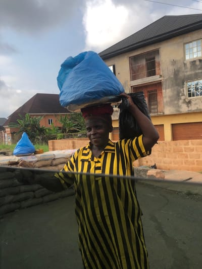 The image depicts an outdoor scene in Ihiala, Nigeria. An adult female stands centrally, wearing a yellow and black vertically striped tunic. Her head is adorned with a reddish fabric wrap, supporting a flat, silver-colored metal tray upon which a large, irregularly shaped object covered in blue plastic is balanced. A black plastic bag is carried over her left shoulder. The woman looks directly towards the viewer with a slight smile.
To her left (viewer's perspective), a smaller, conical object wrapped in blue plastic rests on the ground, positioned next to a stack of multiple "CEMENT" brand sacks. Beyond these sacks, green foliage and bushes are visible.
In the background, two buildings are present. On the right, a two-story structure with light-colored, weathered walls and a dark gray roof is visible. Its upper floor features two windows with security bars, while the ground floor has two closed, orange-brown garage-style doors. To the left, a single-story building with a reddish-brown tiled roof and light-colored walls is partially visible further in the distance. The sky above is overcast with light and dark gray clouds. A dark, horizontal bar or surface extends across the lower portion of the image, suggesting the photograph was taken from within a vehicle. The ground in the immediate foreground appears to be an unpaved, dirty surface.