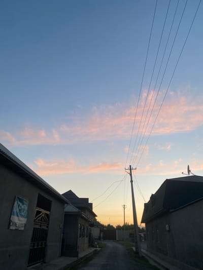 The image depicts a street scene in Боғистон, Tajikistan, during what appears to be sunset or early evening. The sky is a gradient of light blue to pale orange, with wisps of pink clouds scattered across the upper half. Several parallel power lines run diagonally from the upper right to the lower left of the frame. A utility pole with electrical wires is centrally located in the mid-ground, leaning slightly to the left.
On either side of the narrow, unpaved street, there are residential buildings constructed with concrete and grey stucco. The building on the left has a dark-tiled roof and a large metal gate with decorative metalwork. A sign with blue and white lettering is affixed to the wall to the left of the gate. The building on the right also has a dark-colored, sloped roof and a satellite dish mounted on its peak. The architecture suggests a modest, rural or suburban setting. The overall impression is of a quiet, residential street at dusk. There are no people or animals visible in the frame.