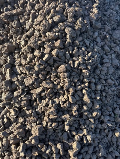 This image displays a large pile of dark, irregular clumps of earth or aggregate material, consistent with excavated soil or crushed construction debris. The material is predominantly dark grey-brown with variations in tone due to direct sunlight illuminating the upper surfaces of many individual clumps, casting distinct shadows towards the lower right within the pile. The texture is rough and uneven, with visible small stones and soil particles compacted into various sizes of irregular lumps. The pile occupies the entire frame, suggesting a close-up perspective or a very large quantity of material. No other objects, people, or discernible background features are present. The scene is located in Broad Chalke, United Kingdom.