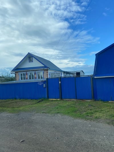 In Malaya Bod'ya, Russia, a two-story house with a blue roof and white siding featuring blue decorative trim is visible. The house has multiple windows with intricate blue and white patterns around them. A bright blue corrugated metal fence runs across the foreground, partially obscuring the house. A street sign on the fence reads "ТРАКТОНАЯ 29" (Tractonaya 29). To the right of the house, another blue corrugated metal structure is visible. The sky above is a mix of blue with scattered white clouds. The ground in front of the fence is covered in gravel and grass.