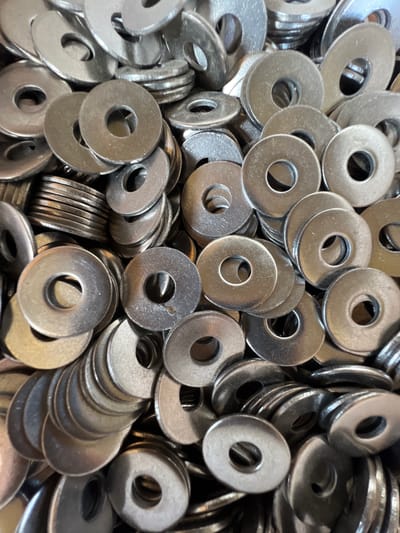The image displays a close-up, overhead view of a large pile of metal washers. The washers are circular with a hole in the center and appear to be made of stainless steel, exhibiting a matte metallic sheen with some reflections. They are stacked and jumbled together, forming a dense, textured surface. Some washers are neatly stacked in small piles, while others are scattered individually. The lighting is somewhat harsh, creating distinct highlights and shadows that emphasize the circular forms and layered arrangement of the washers. The background is filled with more washers, creating a sense of abundance. The image does not contain any contextual information that would allow for its location to be identified as Lanžhot, Czechia.