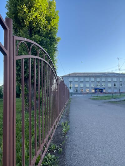 The image shows a long, grey, multi-story building with numerous windows, characteristic of Soviet-era architecture. The building appears to be a residential or administrative block. It is situated adjacent to a paved pathway or road. A brown metal fence with a curved decorative top runs along the edge of the pathway, separating it from an area with green vegetation and trees. The trees are lush and vibrant, suggesting it is daytime in a temperate season. The sky is a clear, bright blue, indicating fair weather. One small, yellow kite can be seen flying in the sky near the top of the building. The overall scene depicts a quiet urban or suburban environment in Kiselyovsk, Russia, likely during the afternoon given the bright sunlight. There is some visible text above the entrance of the building, in blue lettering, but it is not clearly legible.