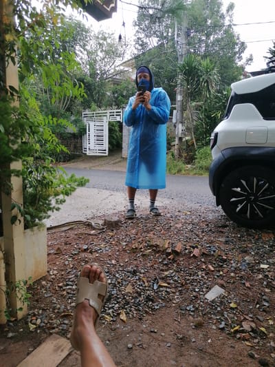The image depicts an outdoor scene with a person wearing a blue rain poncho and a dark blue mask, holding a smartphone to take a selfie. They are standing on a wet, gravelly surface, with a white car parked to their right and lush green foliage in the background. A white gate and a residential building are visible further back. The foreground features a person's leg wearing a light brown sandal, suggesting a low-angle perspective. The environment appears damp, possibly indicating recent rainfall, consistent with the weather conditions often experienced in Lang Biang, Đà Lạt, Vietnam, known for its cool, humid climate and frequent showers. The overall impression is casual and candid, possibly capturing a moment during inclement weather.