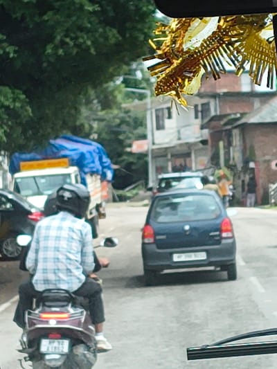 The image shows a street scene in Sidhpur, India, with a focus on two people on a motorcycle. The motorcycle is dark colored with a maroon seat and has a license plate that reads "MP 09 Z 7886". One person is wearing a blue and white plaid shirt and a black helmet, and is seated on the back of the motorcycle. The other person is partially visible, wearing dark pants and light-colored shoes.

Behind the motorcycle, a blue tarp covers what appears to be some goods or equipment. To the left of the motorcycle, a dark blue car is parked on the street. The car has a license plate that is partially visible. Further in the background, buildings with multiple stories are visible, suggesting a semi-urban or town setting. The overall impression is of a street scene, possibly during daylight, with everyday activities taking place. There are trees and greenery in the background, indicating an outdoor setting. A gold, spiky decoration is visible on the left side of the image, partially obscuring the view of the buildings. The image is taken from a slightly elevated perspective, looking down on the scene.