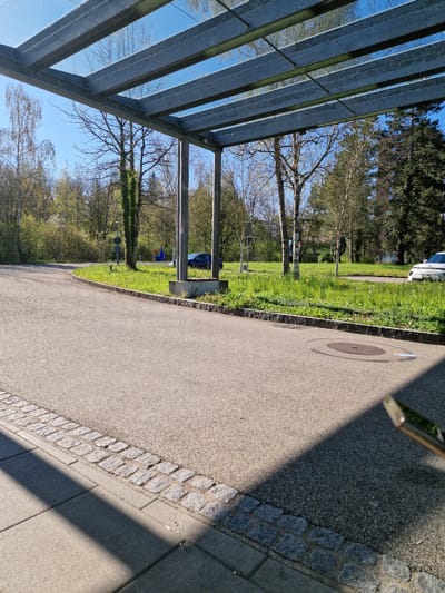 The image shows a sidewalk in Rheinfelden, Switzerland. The sidewalk is made of asphalt with a cobblestone edge. A metal and glass bus shelter is visible on the left side of the image, reflecting the sky and surrounding trees. Adjacent to the sidewalk is a strip of grass with scattered trees and vegetation. Further in the background, more trees and possibly buildings can be seen. A drain cover is visible in the sidewalk.