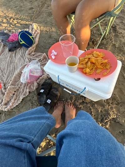 The image shows a close-up view of a beach scene in Castelló de la Plana, Spain, with two people sitting on chairs. The sand is visible, and the lighting suggests it is either early morning or late afternoon.

The foreground is dominated by the legs of two individuals. One person's legs are in the lower right of the frame, wearing blue denim jeans. Their bare feet, with visible toenails and some sand, are also present. The other person's legs are on the left, also bare and showing some tan lines. This person is sitting in a light-colored folding chair.

On the sand, between the two people, are various objects. A white cooler is prominently featured. On top of the cooler, there's a pink plate filled with ruffled potato chips. Next to it, a white cup holds a yellow liquid, likely a beverage. There are also some pink and white heart-shaped candies on a separate pink dish.

Below the cooler, a beige, fuzzy blanket is spread on the sand. On the blanket, a plastic bag contains more pink items, possibly snacks or candy. A blue and yellow disc, possibly a frisbee or a child's toy, is placed on top of some folded clothing, including a pink and white striped item and a grey garment.

Towards the upper right of the image, a pair of black sandals with velcro straps are visible, also covered in sand.

The overall impression is a relaxed beach outing, likely a picnic or a break during a day at the beach. The presence of snacks, drinks, and leisure items suggests a casual and enjoyable experience. The text "La Nevera" is visible on the side of the white cooler.