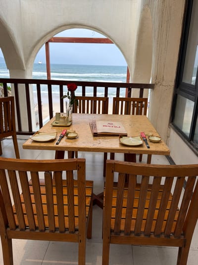 An outdoor dining area in An Hải Ward, Vietnam, features a square, light-colored table with a dark wooden base. The table is set for two, with each place setting comprising a square, patterned plate, a small circular dish, and chopsticks in a red wrapper. A stack of red-covered menus rests near the center, alongside a small tray holding a creamer and sugar bowl. A single red rose is displayed in a clear glass vase. Six wooden chairs with slatted backs and seats are arranged around and near the table. The dining space is framed by off-white arched architecture and separated from a sandy beach by a wooden railing. Beyond the beach, the ocean displays gentle waves under an overcast sky. A large vessel is visible on the distant horizon, with landmass further to the left. No individuals are present in the scene.