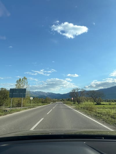 The image captures a picturesque road scene from the perspective of a vehicle, likely during a journey through Mezdreya, Bulgaria. An asphalt road, marked with white dashed and solid lines, stretches diagonally into the distance, bordered on the right by a darker section of the car's interior.
To the left of the road, lush green fields transition into a landscape dotted with deciduous trees, some of which appear bare or just beginning to show new leaves, suggesting late winter or early spring. A prominent range of mountains dominates the background, rising under a clear, bright blue sky filled with scattered white cumulus clouds. Sunlight illuminates the scene, casting a clear, bright glow.
Along the roadside, two signs are visible on the left: a round white sign with a red border, and closer to the foreground, a larger, dark rectangular sign whose text is not readable due to distance. The overall impression is one of a serene drive through a rural or mountainous area on a sunny day.