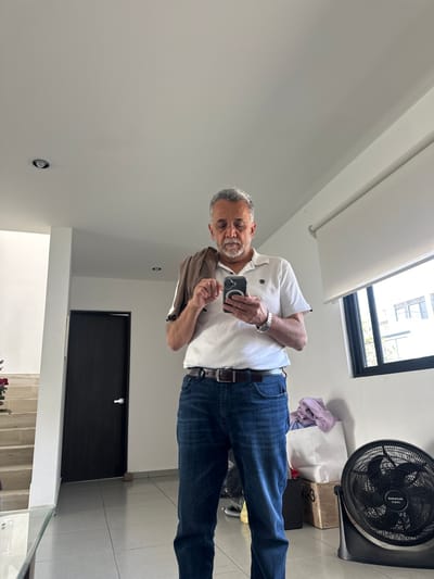 An older man in jeans and a white collared shirt is taking a selfie in a well-lit room. He is holding a phone in his right hand. In the background, there's a black door, a ceiling fan, a skylight, and what appears to be the top of a staircase with plants. The room has white walls and tile flooring. A brown jacket is draped over his left arm.