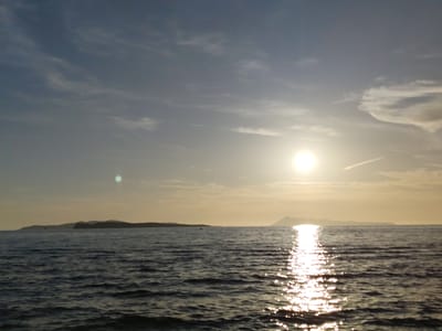 This image captures a seascape at either sunrise or sunset in Agios Stefanos, Greece. The horizon line is positioned approximately at the image's midpoint, dividing the frame into a sky upper half and a sea lower half.
The sky exhibits a gradient of muted blue-gray tones, transitioning to a pale yellow-orange near the horizon due to the sun's presence. Scattered, wispy white clouds are visible across the sky. A bright, circular sun is prominent in the mid-right portion of the sky, casting a strong glare. A distinct green circular lens flare is present in the upper left quadrant, and a faint contrail crosses the sky in the upper right.
The sea in the foreground is dark blue-gray with a rippled, textured surface indicating small waves. A brilliant, shimmering path of sunlight reflects across the water, originating from the sun and extending towards the viewer, creating numerous bright specular highlights on the water's surface.
Two distinct landmasses are visible on the distant horizon. The landmass to the left is lower and extends horizontally, while the one to the right appears higher and more mountainous, both silhouetted against the brighter sky. An unidentifiable small, dark object is faintly discernible on the water's surface between the left landmass and the sun's reflection path. No people, discernible boats, or other prominent man-made structures are present in the foreground or midground.