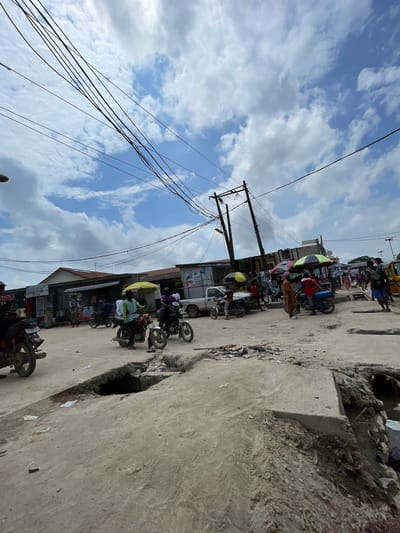 The image depicts a bustling street scene in Lagos, Nigeria, with a prominent focus on the urban environment and the daily activities of its inhabitants. The foreground is dominated by a cracked and uneven concrete road, with a large, dark opening suggesting a damaged drain or sinkhole. Debris is scattered across the ground, indicative of ongoing development or wear and tear.
In the mid-ground, motorcycles, a common mode of transport in Lagos, are parked and in motion. Several individuals are seen riding or standing near their motorcycles. Various shops and stalls line the street, with awnings and umbrellas providing shade. Some of these establishments display signage, although the text is largely illegible. People are casually dressed and engaged in various activities, suggesting a marketplace or commercial area. The presence of umbrellas, some with bright yellow and green colors, adds a splash of vibrancy to the scene.
The background reveals more buildings, utility poles with a dense network of overhead cables, and a truck. The sky is a mix of blue with scattered white clouds, indicating daylight. The overall impression is one of a dynamic and active urban setting, characteristic of many parts of Lagos, Nigeria. The scene is candid and captures the raw, unadulterated essence of street life in the city.