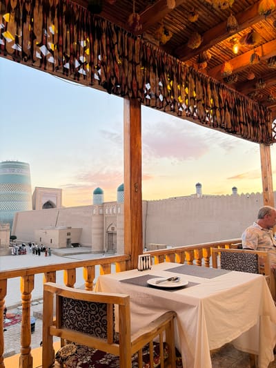 The image depicts a table set for two on a wooden balcony overlooking the city of Khiva, Uzbekistan. The balcony is adorned with decorative wooden railings and a rustic wooden ceiling from which woven tassels and a light bulb hang. A white tablecloth covers the table, which is set with a plate, silverware, and a napkin holder containing forks and knives. A man, partially visible, sits at the table to the right. In the background, the historic architecture of Khiva is visible, including the Kalta Minor minaret, various mosques with turquoise domes, and ancient mud-brick walls. People are seen walking in the courtyard below. The sky is a soft gradient of orange and blue, suggesting it is sunset.