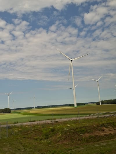 The image shows a field with several wind turbines under a cloudy blue sky. The wind turbines are white and tall, with three blades each, and appear to be operational. The foreground of the image consists of green grass and a wire fence. In the middle ground, there are fields of crops in various shades of green and yellow, suggesting agricultural land. In the background, there is a line of trees and a distant horizon. The overall scene depicts a rural landscape with renewable energy infrastructure. This scene is located in the village of Zabiele, Poland.