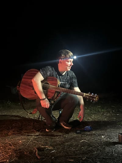 The image shows a man seated outdoors in a dark environment, possibly Юзуфоўскі сельскі Савет, Belarus. He is wearing a headlamp, a patterned t-shirt, dark pants, and sneakers. He is holding and playing an acoustic guitar. To his right on the ground are a blue object, a small black device, and a metal canister. The ground is covered with grass, leaves, and twigs. The overall impression is of a person playing music in a dimly lit, natural setting.