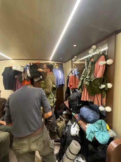 Two men are packing or unpacking items in a small, cluttered room. The room appears to be a storage space or closet. Garments and backpacks hang from hooks on the top of the wall. One man wears a gray shirt. The other man wears glasses and a green long-sleeved shirt.
The location cannot be determined as Mira-Bhayander, India from the image.