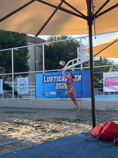 The image depicts a beach volleyball scene in Gošići, Montenegro. A female athlete is captured in mid-air, executing a volleyball spike or serve on a sandy court. She is wearing a purple and pink sports bra and white bikini bottoms. A volleyball is positioned above her right hand, indicating the moment of impact or preparation for a hit.
Behind the court, a blue banner with white text reads "LUŠTICA 2023" and other smaller text and logos, suggesting it's a promotional banner for an event or location. To the right of this banner, a white banner with pink lettering displays "ELITE VOLLEYBALL". A chain-link fence runs along the perimeter of the court, separating it from a grassy area and buildings in the background.
The environment is outdoors, under a clear sky, with the warm light of what appears to be late afternoon or early morning. Several large, beige umbrellas with dark frames provide shade. The sandy ground shows footprints and the texture of disturbed sand. In the foreground, a blue mat is visible, with a red bag and a pair of sunglasses lying on it. The overall setting is that of a beach sports facility.