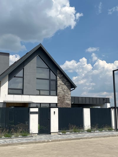 The image depicts a modern, two-story house with a dark gray pitched roof and large triangular windows on the upper floor. The exterior walls are a mix of white stucco and brickwork. A dark gray metal fence with vertical slats runs across the front of the property, with a gate in the center. White pillars are integrated into the fence design. The foreground shows a paved walkway and a street. The sky above is blue with scattered white clouds. The house is located in Strumivka, Ukraine, as indicated by the inscription on one of the white pillars which reads "Струмівка, 4". There are no people or animals visible in the image.