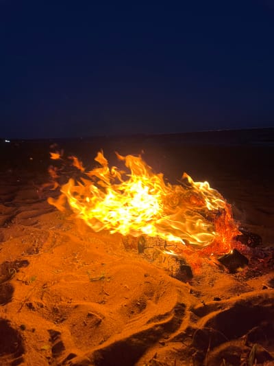 The image depicts a campfire burning on a sandy beach at night. The fire is the central subject, with bright orange and yellow flames consuming logs. The sand surrounding the fire is illuminated by its glow, casting long shadows and highlighting the texture of the sand. In the background, the dark expanse of the sky meets a faint horizon line, possibly indicating a body of water and distant lights. The overall atmosphere is one of warmth and tranquility in a natural setting. The location is identified as the city of Solnechnoye, Russia.