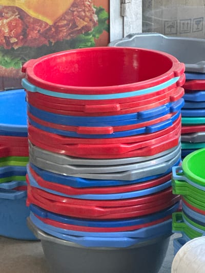This image captures a close-up view of a stack of colorful plastic buckets. The main focus is on a large red bucket at the top of a tall, neatly organized pile of buckets in various shades of red, blue, and gray. To the left, a portion of a large blue bucket is visible. A smaller stack of green and blue buckets is on the right side of the frame. The background features a blurry advertisement for a fast-food item, possibly a burger, with a white building structure visible behind it. The scene is illuminated by natural light, casting soft shadows. The setting appears to be an outdoor market or a retail space where these buckets are being sold. The exact location in Kara-Suu, Kyrgyzstan, is not explicitly identifiable from the visual cues alone, but the presence of a market or shop is implied. There are no people or animals visible in the image.