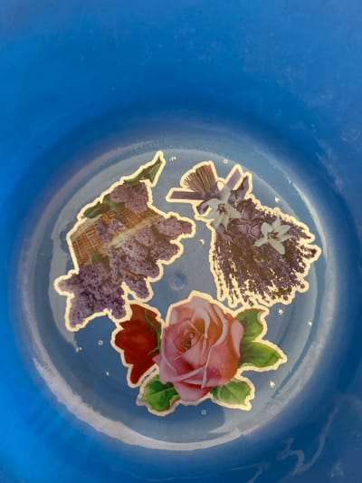 This is a top-down, close-up shot of a blue plastic bowl's interior, which has a subtle, glossy sheen, suggesting it might be wet or recently cleaned. The bowl is decorated with three stickers of flowers, outlined in gold. The stickers are slightly peeling at the edges.
The subjects are the floral stickers. One sticker depicts a bouquet of lavender with white flowers and a purple ribbon tied around it. Another shows a pink rose with green leaves and a hint of a red rosebud below it. The third sticker features a cluster of purple lilacs in a woven basket. The stickers appear to be applied to a light blue surface within the bowl, which is itself a deeper shade of blue. Small, scattered white dots, resembling stars or glitter, are also visible on this lighter blue surface, adding a decorative element.
There is no activity or event taking place. The image captures a static scene.
Notable details include the textured appearance of the flower stickers, the subtle shine of the plastic bowl, and the decorative elements of the white dots. The lighting appears to be bright, casting soft reflections on the bowl's surface. The location is identified as Bulamare, Nigeria, but there are no visual cues in the image itself that directly indicate this specific location; the context is provided externally.
There is no visible text in the image.