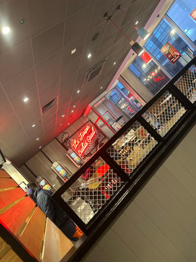 This image depicts the interior of a KFC restaurant located in Chatham, United Kingdom, captured from an elevated, angled perspective looking downwards into the main dining area and towards an exterior street view.
In the foreground, on the left, a wooden staircase with illuminated red-orange underside lighting ascends. A single individual, wearing a dark blue jacket with visible orange accents on the lower back, is partially observed on the lower steps of this staircase, facing away from the camera. A black metal railing with a diamond mesh infill panel extends diagonally across the mid-ground, separating the elevated viewpoint from the restaurant floor below.
Beyond the railing, the primary restaurant area features a modern design. Two interactive touchscreen self-service ordering kiosks displaying food items are visible against a light wood-paneled wall. Above and to the right of the kiosks, a large red neon sign spells "It's Finger Lickin' Good" and incorporates a stylized chicken graphic. Further into the restaurant, multiple tables and chairs, some with red and yellow elements, are arranged, occupied by several indistinct patrons.
The ceiling consists of grey acoustic tiles with recessed lighting fixtures and an exposed HVAC vent. Three pendant lights with cylindrical shades (one light blue, one black, one red) hang from red cables from the ceiling towards the upper right. Large glass windows on the far right provide a view of an urban street or public area, with reflections of buildings and distant signage visible. The exterior environment appears to be an urban commercial area.