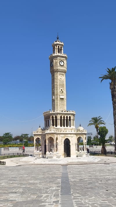 This media file captures a view of the Izmir Clock Tower, a prominent landmark in Izmir, Turkey. The tower stands tall against a clear, bright blue sky, suggesting a sunny day. It is made of light-colored stone with intricate carvings and decorative details. The clock face is visible on the tower, indicating a daytime setting.

At the base of the tower is an ornate structure with arches and columns, and a dark, decorative gate. In the foreground, the ground is paved with grey cobblestones. To the right of the tower, a tall palm tree stands, its fronds rustling slightly. In the background, there are more palm trees, lush green foliage, and a body of water, hinting at a coastal or park-like setting.

There are a few people visible in the scene. One person in a red shirt is walking in the middle ground to the left of the tower. Other individuals can be seen seated on benches in the background. The overall atmosphere appears peaceful and tranquil, as indicated by the calm weather and the presence of people enjoying the public space. The scene is likely taking place in the morning or early afternoon, given the bright sunlight and clear sky. No visible text is present in the image.