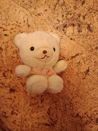 A white teddy bear is placed on a textured, wooden surface. The teddy bear is plush and has black button eyes, a brown nose, and a pink stitched mouth. It is wearing a pink, lacy ribbon around its neck. The wooden surface has a warm, orange-brown color with dark speckles and irregular lines, suggesting a distressed or natural wood grain. The lighting casts subtle shadows, highlighting the texture of the wood and the softness of the bear. The scene is depicted from a top-down perspective. This image is not specific to Naro-Fominsk, Russia, or any other location.