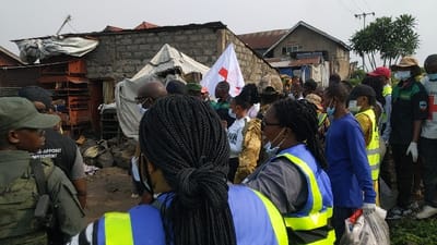 The image displays an outdoor scene in Gisenyi, Rwanda, featuring approximately two dozen individuals gathered on what appears to be an unpaved thoroughfare amidst an urban environment. Many participants, predominantly adults, are wearing face masks. Several individuals are attired in high-visibility vests: some in blue with bright yellow accents, others in dark green. One person wears a military-style camouflage uniform and cap. A white flag bearing a prominent red cross symbol is visible. On the right, an individual wears a dark shirt with a Red Cross emblem, white gloves, and holds a clear plastic bag containing items. The background features multiple single and two-story structures constructed from unpainted cinder blocks with corrugated metal roofs. Some rooftops are partially covered with tarpaulins. Debris, including rocks and miscellaneous items, is scattered on the ground. An antenna is mounted on one roof. The sky is overcast.