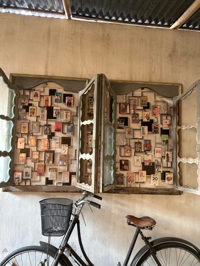 The image depicts an interior scene in Banguntapan, Indonesia, featuring a wall adorned with an eclectic display of cassette tapes housed within decorative frames, set against a backdrop of vintage-style wallpaper. Two open, antique wooden window frames serve as the primary display units, showcasing rows of cassette tapes arranged in a grid-like fashion. The cassette tapes, many of which appear to be music albums, are diverse in their cover art and titles, suggesting a curated collection. Some notable titles visible include "Michael Jackson," "ABBA," and "Car."

In the foreground, a vintage bicycle is positioned to the left of the frame, facing towards the right. The bicycle is dark in color and equipped with a black wire mesh basket mounted on the handlebars, which is empty. Another bicycle, partially visible on the right side of the image, is also dark and features a brown leather saddle. The wall itself is textured and aged, with visible cracks and imperfections, contributing to the rustic ambiance. Above the display, a corrugated metal roof with visible supporting beams is present, suggesting an outdoor or semi-outdoor covered area. The overall scene evokes a sense of nostalgia and artistic arrangement, likely in a cafe, shop, or private residence in Banguntapan.