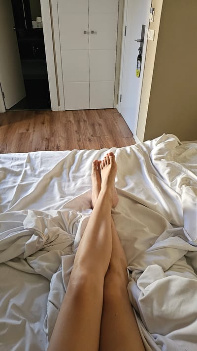 A first-person perspective shot captures a person's legs and feet resting on a rumpled white duvet in a bedroom. The feet, with visible toes and toenails, are crossed at the ankles. The legs are tanned and smooth. The bed is situated on a wooden floor, with a section of closet doors and a white door visible in the background. The closet doors are a plain white, with two square handles on each of the three panels. The white door has a silver handle and a black tag hanging from it. The room appears to be in a hotel, with the tag likely a "Do Not Disturb" sign. The lighting suggests it's daytime, with sunlight casting shadows on the wooden floor and the wall. The environment is tidy and uncluttered.