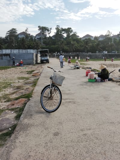 A bicycle with a wire basket is parked on a concrete path. To the left of the path is a sandy area with some sparse vegetation and rocks. In the background, a low, corrugated metal wall separates the sandy area from a street with several parked cars and a small truck. Beyond the street, lush green trees and palm trees are visible, with several modern houses situated on a hill. Further in the background, the sky is a pale blue with scattered white clouds. Several people are seen walking along the street or near the sandy area. In the mid-ground on the right side of the path, a person wearing a conical hat is sitting near a stack of containers filled with food and a red bag. Another person is sitting behind a table on the left side of the path. The scene appears to be a public area near the coast in Phú Thủy, Vietnam.