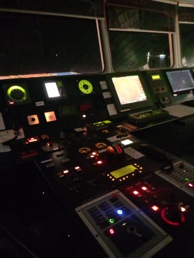 The image shows the control panel of a ship's bridge at night. Various screens, dials, and buttons are visible, some illuminated with green and red lights. The displays show what appear to be navigation and operational data. The overall setting is dark, suggesting the image was taken in low-light conditions, possibly during nighttime operations. The environment appears to be an indoor, technical space within a vessel. There are no people or animals visible in the frame. The location is unknown and it is not possible to determine if it is in the city of Unknown, United Kingdom from the image.