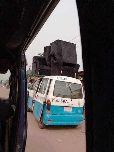The image captures a street scene in Orie Uratta, Nigeria, viewed from within another vehicle, with its dark interior framing the left and right edges. A white and light blue commercial van, marked with "ICT/MCCRD" and "IM00460A" on its rear, is central. Two dark, upholstered armchairs with gold-colored leg accents are stacked on the van's roof. The van's rear passenger door is open, extending outwards. Its license plate holder displays "01U-162." The road is unpaved, consisting of reddish-brown dirt with scattered debris. In the blurry background to the left, other vehicles, potentially motorcycles, and indistinct figures of people are visible, suggesting activity on a thoroughfare. Buildings with corrugated roofs and some sparse foliage are present along the street. The sky is uniformly overcast.