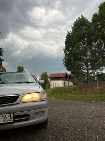 The image depicts a silver car parked on a gravel road. The car's headlights are on. In the background, there is a house with a dark roof and a white base. There are trees and a cloudy sky. The license plate on the car reads "161 RUS". This scene is located in Tomsk, Russia.