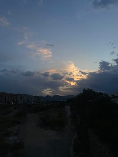 The image depicts a twilight scene in Sagunto, Spain, with a dramatic sky filled with dark, brooding clouds contrasted by streaks of pale yellow and pink light near the horizon. In the mid-ground, a low-lying urban landscape with buildings lines the base of hazy, dark mountains. The foreground is dominated by a dimly lit, unpaved path or dirt road winding through dry, sparse vegetation. To the right, a dense area of dark foliage partially obscures a stone wall. The overall atmosphere is somber and quiet, suggestive of the end of the day.