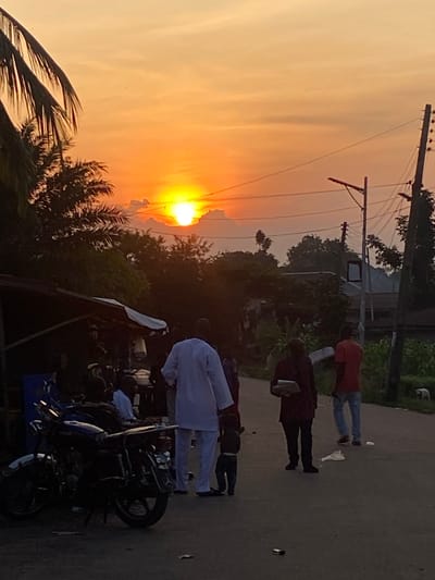 This image captures a sunset scene in Nguzu Edda, Nigeria, depicting a bustling street environment during the golden hour. The sky is dominated by intense orange and yellow hues, with the sun prominently visible on the horizon, partially obscured by dark cloud formations. Dense tropical vegetation, including large palm fronds on the left and various trees and bushes, is silhouetted against the bright sky, primarily on the left and in the background.
On the left side of the frame, a makeshift market stall or small shop with a dark roof and a white canopy is visible, where several indistinct figures are gathered. Parked on the road near this stall is a dark-colored motorcycle, likely blue or black, featuring chrome fenders and a visible license plate frame, oriented towards the right.
The central and right portions of the image feature a dark, likely asphalt or packed dirt road. Along the right side of the road, multiple wooden utility poles with an array of power lines and a street light fixture extend vertically.
Several individuals are present on the road, walking towards the right. In the midground, a person wearing a white, traditional long-sleeved garment and trousers is walking, accompanied by a small child in dark clothing walking beside or slightly behind them. To their right, another individual in dark clothing is walking while holding a rectangular object resembling a tray or container. Further right, a person in a red shirt and blue jeans is also walking away from the viewer. Scattered debris, including a crumpled white object and a small dark item, is visible on the road surface. In the far right midground, a small, light-colored dog is observable near the edge of the road. Distant structures and more vegetation are faintly visible further down the street on the right.