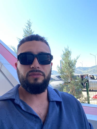 A man with dark hair and a beard wearing black sunglasses and a dark blue shirt with a repeating pattern is taking a selfie outdoors. He is positioned in the foreground, with a bright blue sky and distant mountains in the background. Several evergreen trees are visible, along with parts of vehicles and a light-colored structure with pink and white stripes. The scene appears to be in a somewhat arid, possibly mountainous region. The man is looking directly at the camera.