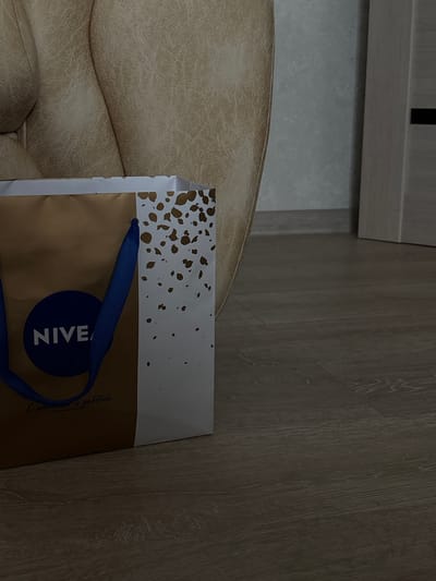 The image presents an indoor scene featuring a NIVEA shopping bag next to upholstered furniture and wooden flooring. No people are visible.

The main subject is a NIVEA branded shopping bag, colored gold/brown with a white top border decorated with scattered golden, irregular speckles. It features blue ribbon handles and a prominent dark blue circular NIVEA logo. Below the logo, faint Cyrillic text is visible, reading "С любовью и заботой," which translates to "With love and care." The bag is positioned partially resting on a light brown or beige upholstered piece of furniture, likely an armrest or backrest of a chair or sofa, which occupies the lower-left portion of the frame and shows a textured surface.

The setting appears to be a room, possibly a home, in Zhemkovka, Russia. The floor is covered with light-colored wooden or laminate planks, displaying distinct grain patterns. In the upper left corner, the base of a light-toned wooden cabinet or piece of furniture is visible against a plain, light grey wall or surface. The lighting is soft and ambient, suggesting an indoor shot during an indeterminate time of day. There is no specific activity taking place; the bag simply rests in the scene. No particular emotions are conveyed.
