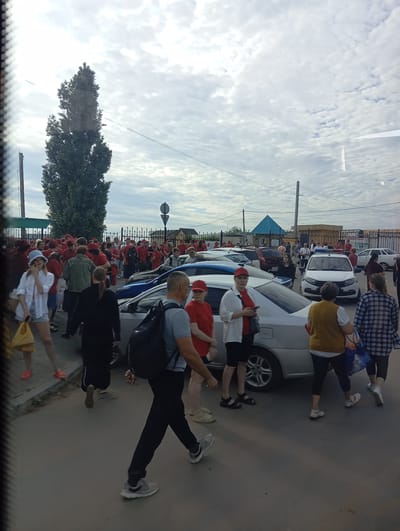 The image depicts a scene outside a building in Sol-Iletsk, Russia, under a cloudy sky. A crowd of people, many wearing red clothing and caps, are gathered in an outdoor area that appears to be a parking lot or street. Several cars are parked and visible, including a white police car with flashing lights. Some individuals are walking, while others are standing and interacting. The overall atmosphere suggests an organized gathering or event. The presence of a tall, slender tree and a fence with decorative elements are notable environmental details.