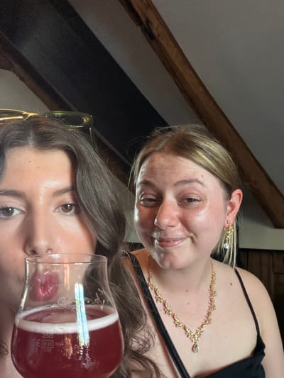 The image is a selfie of two young women with drinks. The woman on the left is in the foreground, holding a glass of red liquid with white foam. Her face is partially obscured by the glass, but her eyes and lips are visible. She has dark, wavy hair and is wearing sunglasses on her head. The woman on the right is smiling and looking at the camera. She has blonde hair and is wearing a gold necklace with a pendant and dangling earrings. She is wearing a black tank top. The background appears to be an indoor setting with wooden beams visible on the ceiling. The location is not identifiable as Longueuil, Canada.