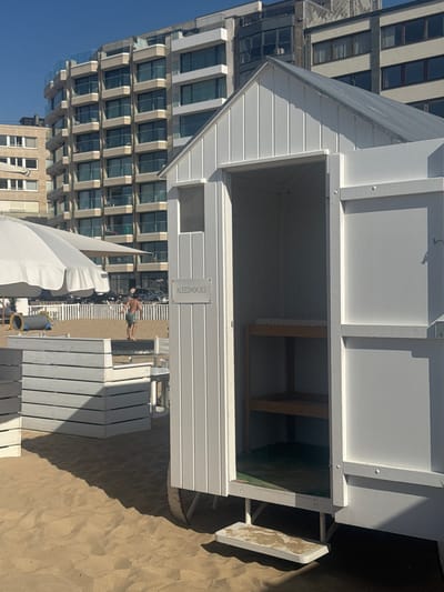 The image shows a beach scene in Knokke-Heist, Belgium. A white beach changing hut with a sign reading "KLEEDHOKJES" is prominently featured. Inside the hut, a wooden shelf is visible. Outside, a row of white beach chairs stretches towards the horizon. A shirtless man in swimming trunks is walking in the distance. A partially visible beach umbrella is in the foreground. Behind the changing hut and beach chairs is a tall modern building with numerous windows, typical of the architecture in Knokke-Heist. The ground is sandy. The sky is clear and blue.