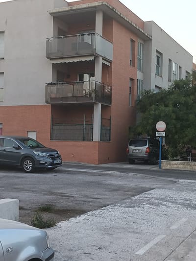The image shows a multi-story apartment building with red brick and light-colored facade elements. The building has several balconies with glass railings. In front of the building, on the street, are two parked cars: a dark grey SUV on the left, and a silver minivan on the right. A blue pole with a circular red and white sign indicating "EXCEPTE VEINS" (Except residents) stands near the silver minivan. The street is paved and there is some vegetation and a low stone wall in the background. The sky is overcast. The image appears to have been taken from a street-level perspective. This scene is consistent with a residential street in Tortosa, Spain.