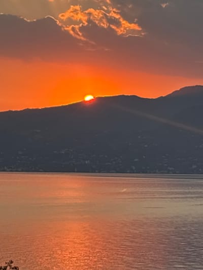 The image depicts a sunset over Lake Garda, Italy. A sliver of the sun is visible, peeking over the silhouette of a mountain that dominates the left side of the frame. The sky is filled with fiery orange and yellow hues, with some wispy clouds catching the last rays of sunlight. The right side of the image is a calm body of water, reflecting the vibrant colors of the sky. The water appears to have small ripples, creating a textured effect. The overall mood of the image is serene and picturesque, capturing the beauty of a sunset in a natural landscape. There is no visible text in the image.