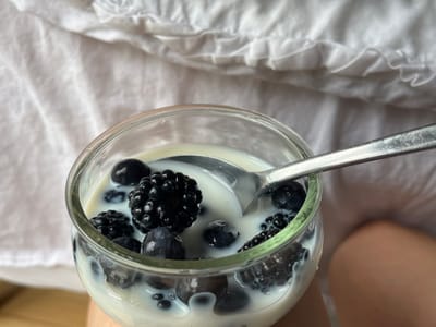 The media file presents a close-up, high-angle shot of a glass bowl filled with blackberries and blueberries submerged in white milk or yogurt. A silver spoon rests within the mixture, partially covered by the liquid and berries, suggesting it's either just about to be eaten or has just been stirred.
The most prominent subjects are the berries: several large, textured blackberries and smoother, smaller blueberries are clearly visible. They are a deep, rich blue-black, contrasting with the creamy white liquid. The glass bowl is simple and clear, allowing the contents to be fully seen. The spoon is metallic and shows some reflections.
The background consists of soft, white fabric with visible wrinkles and folds, possibly bedding or a blanket, giving the scene a cozy and intimate feel. In the bottom right corner, a segment of a bare human leg or knee is visible, hinting that the person is holding or resting the bowl on their lap.
The activity taking place is the consumption of a light meal or snack, likely breakfast or a refreshing treat. The setting appears to be indoors, potentially a bedroom or a comfortable living space, emphasizing a moment of personal enjoyment and relaxation in Aarhus, Denmark.
Notable details include the natural light illuminating the scene, creating soft shadows and highlights on the berries and spoon, which suggests it could be morning or early afternoon. The overall impression is one of simplicity, freshness, and comfort. There is no visible text in the image.