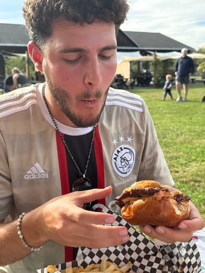A young adult male, with curly brown hair and a trimmed beard, is the primary subject, shown with his eyes closed and mouth slightly open, appearing to be in the process of eating. He wears a light tan Adidas short-sleeve shirt with red and black vertical stripes and the "AJAX AMSTERDAM DAILY PAPER" logo, including three stars, on the right chest. A dark beaded necklace with a dark pendant is around his neck, and a light-colored beaded bracelet is on his left wrist. He holds a partially consumed burger, featuring two meat patties, sauce, and cheese within a brioche bun, with both hands. A black and white checkered paper liner is visible beneath the burger, and french fries are in a container in the lower left foreground. The scene is an outdoor grassy area located in Accord, United States. In the background, a building with a dark roof is visible, along with multiple white canopy tents providing shaded seating. Several blurred individuals are present: a person in a dark top on the far left, a man in a dark top, light shorts, and a cap standing, and a small child in a blue outfit walking on the grass. The sky is partly visible overhead.