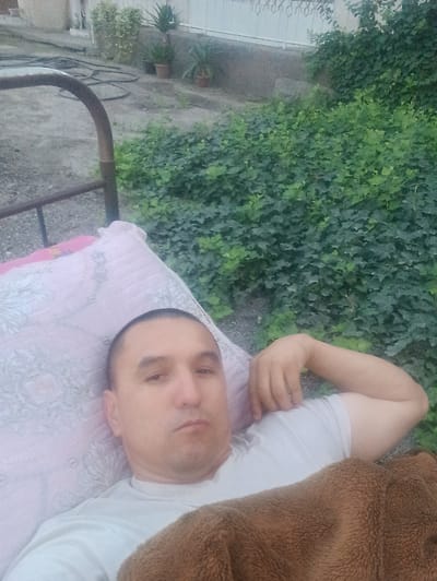 In Chumbagish, Uzbekistan, an adult male is captured in a selfie lying on a bed with a patterned pink pillow. He is wearing a white t-shirt and is partially covered by a brown blanket. The background reveals a dense green foliage of ivy plants against a white fence and some potted plants. The ground appears to be gravel and dirt, with a black hose visible. The scene suggests an outdoor setting, possibly a backyard or patio area.