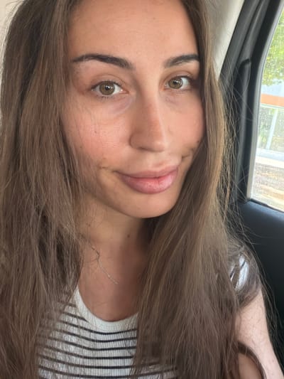 A close-up, head-and-shoulders selfie of a woman with long, brown hair is taken from a slightly upward angle, likely inside a vehicle. The woman has fair skin, brown eyes, and a subtle smile. She is wearing a striped shirt and a delicate silver necklace. The background shows a car window with a blurred view of trees and a building outside, suggesting an urban or suburban setting in Larnaca, Cyprus. The lighting is natural and appears to be daytime.