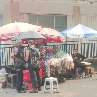 This image captures an outdoor street vendor setup in the Special Capital Region of Jakarta, Indonesia. The scene is illuminated by bright daylight.
In the foreground, a mobile vendor cart stands on worn asphalt pavement. The cart is laden with numerous packaged snacks and bottled beverages, prominently displayed. Overhead, two large red umbrellas are visible, both bearing the "Pucuk Harum" brand logo, consistent with a popular Indonesian tea. A smaller, plain black umbrella is also present. To the right of the vendor cart, a blue and white umbrella with the "Biznet" logo, an Indonesian internet service provider, provides additional shade.
Three male individuals are present. On the left, an older man with grey hair, wearing a grey short-sleeved top over a dark long-sleeved shirt, black cargo pants, and black boots, stands with his left hand on his hip. He is facing and seemingly conversing with a second man, who is also standing and dressed in dark clothing, black pants, and black boots, with a camouflage-patterned backpack. This second man has his back partially turned to the viewer. A red Coca-Cola branded cooler is situated between these two men and the vendor cart.
To the right of the vendor cart, a younger man, dressed in a black long-sleeved shirt and dark pants, is seated on a light-colored stool. He holds a dark smartphone in his right hand and appears to be looking forward. A black bag is draped near him, and a small brown plastic stool is positioned close by.
In the background, a black metal fence or railing separates the street from a light-colored building with large windows. An empty white plastic stool is visible in the immediate foreground.